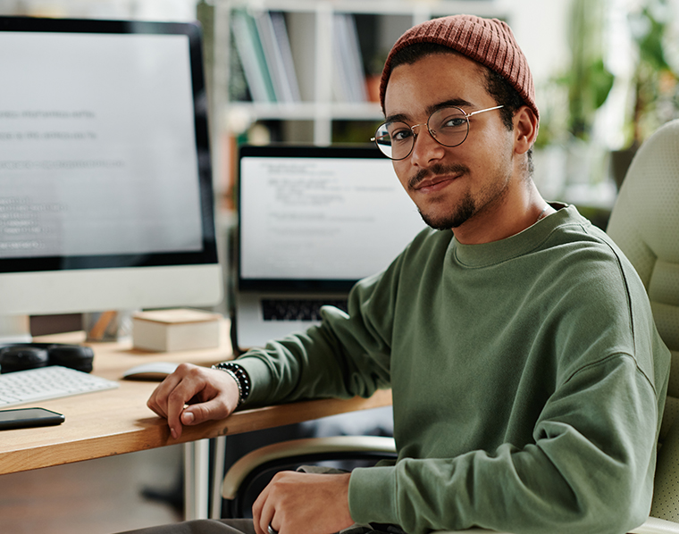 Young confident IT engineer or software developer in casualwear sitting in armchair by computer monitor and laptop and looking at camera