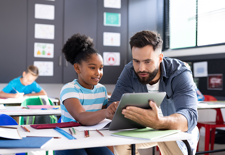 Diverse male teacher and happy girl using tablet at her desk in elementary classroom